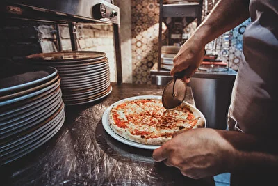 Pizzaiolo che taglia con la mezzaluna una pizza margherita appena sfornata, accanto a una pila di piatti