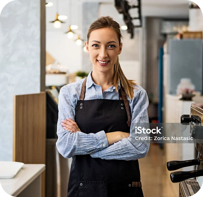 Titolare di un'attività con grembiule da lavoro sorridente al bancone, tra vetrina dei dolci e macchina del caffè